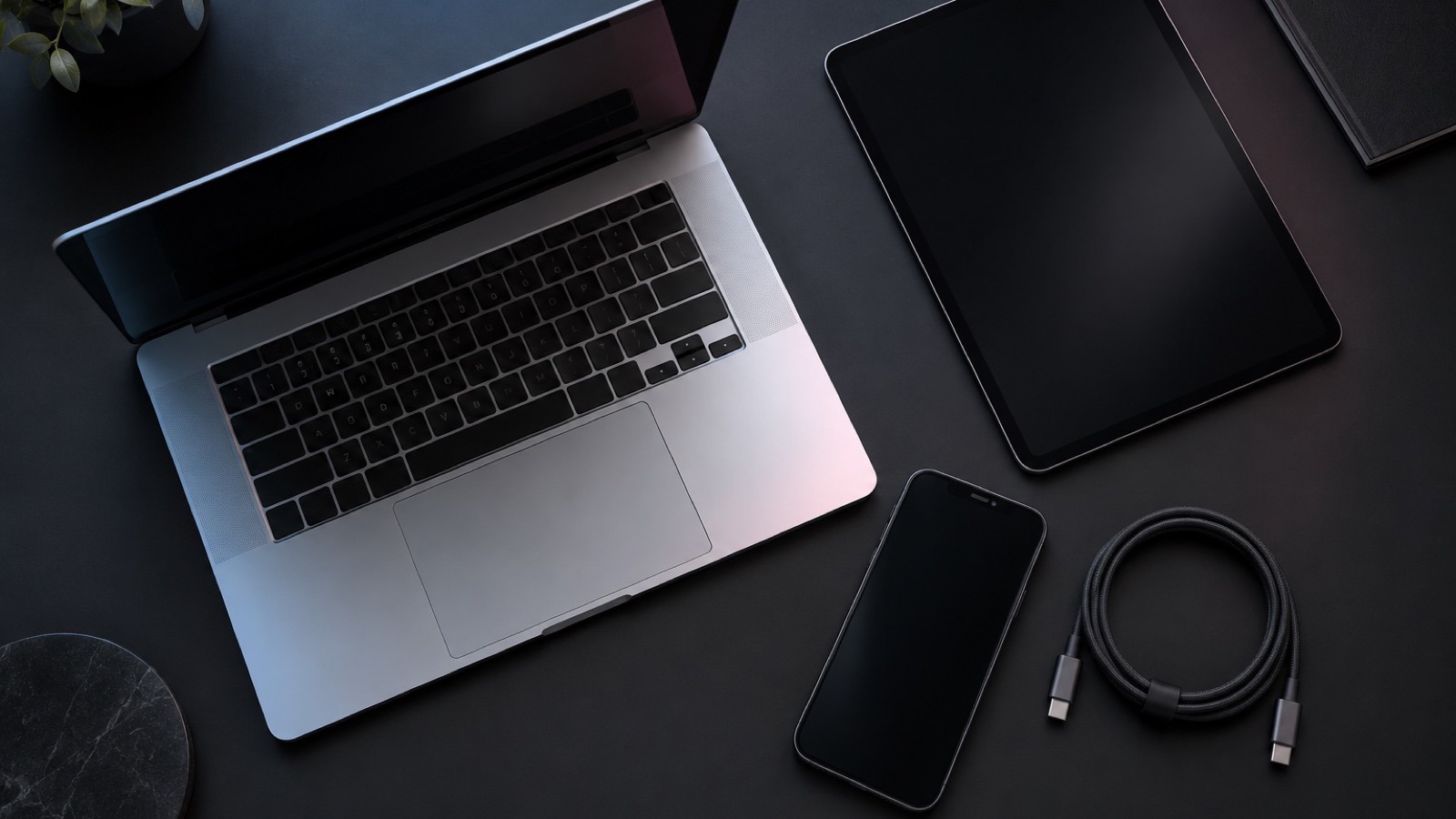 A silver laptop, graphite tablet, and smartphone with a charging cable on a charcoal work surface.