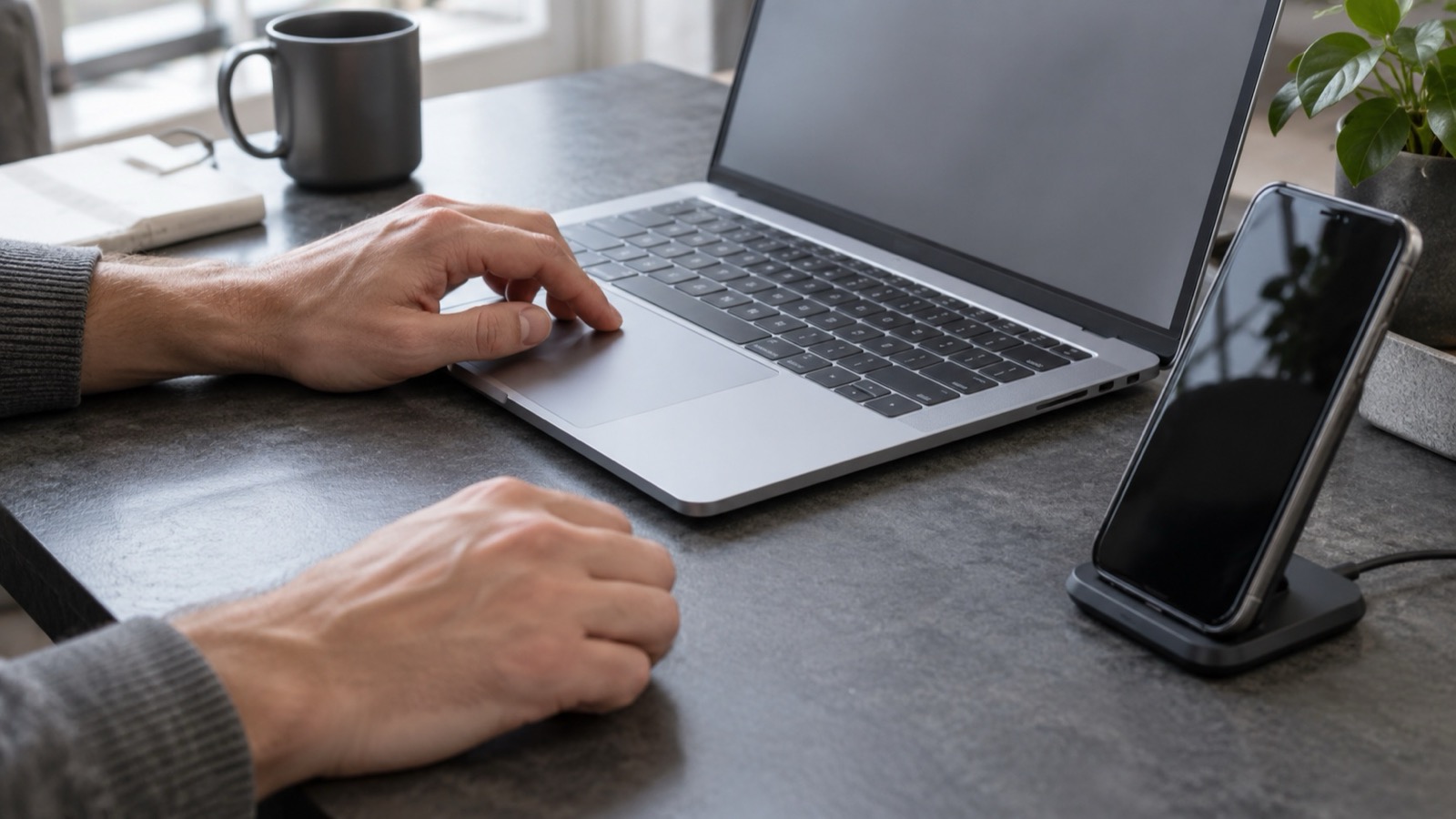 Hands using a silver laptop beside a graphite phone on a charging stand in a daylight workspace.