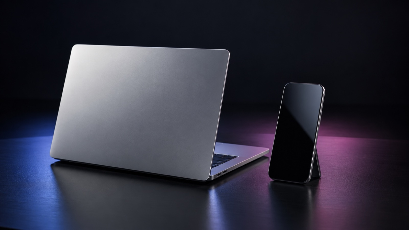 An open silver laptop and graphite phone on a black tabletop with subtle blue and pink reflections.