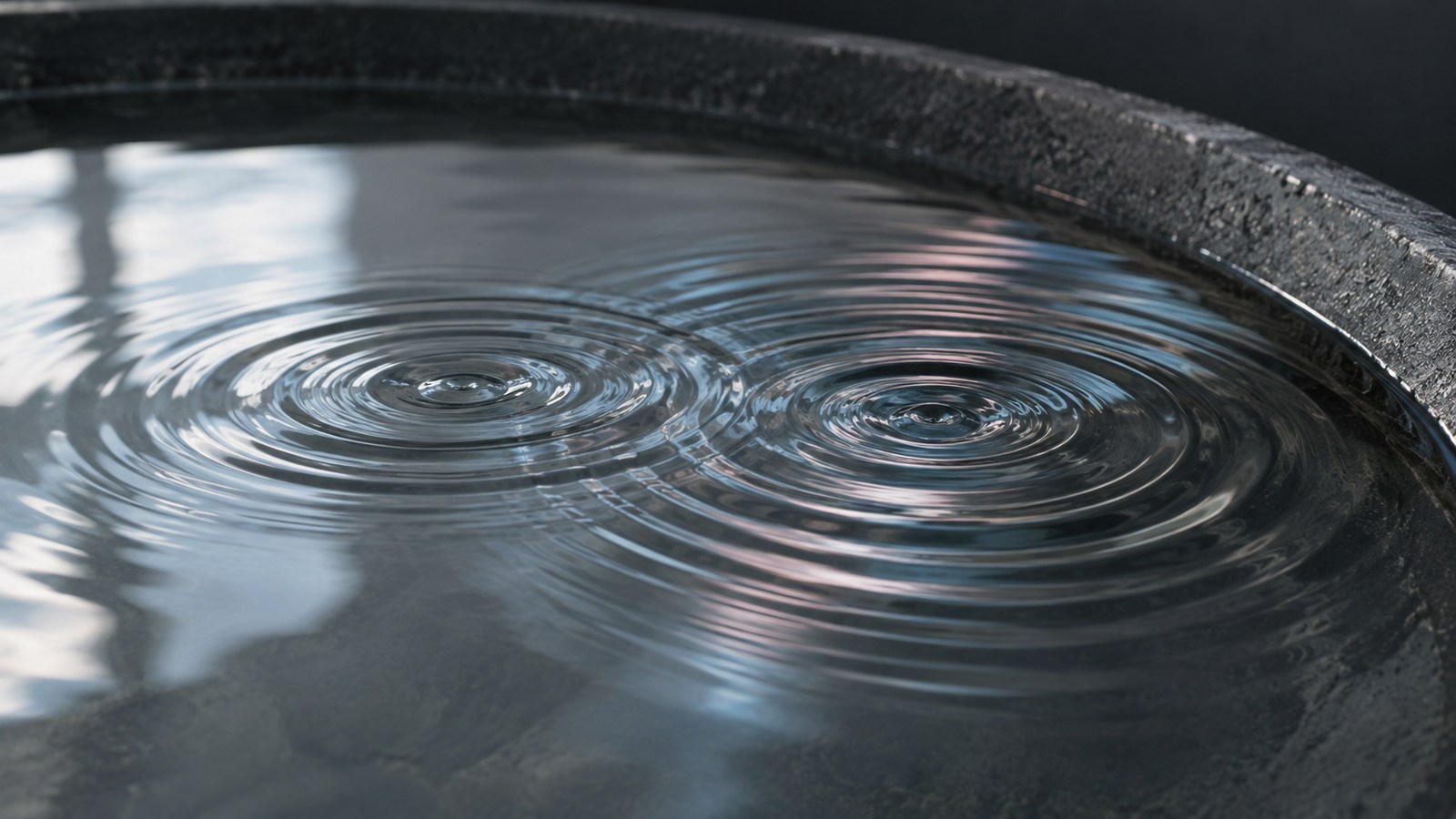 Intersecting ripples in clear water inside a shallow graphite stone basin.
